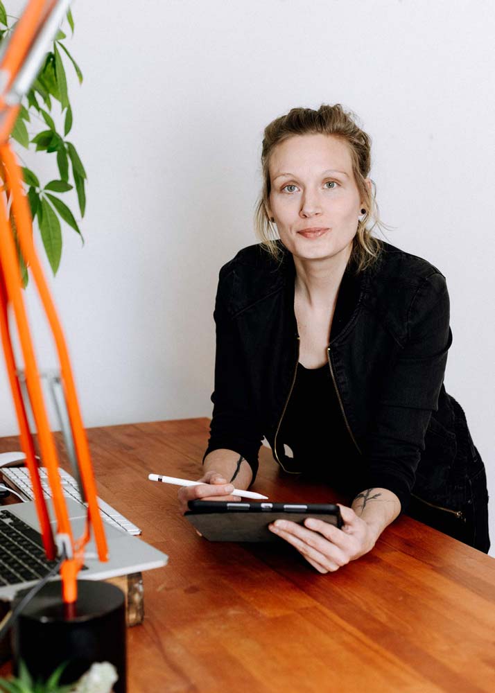 Kathrin Windhorst sitzt an einem Schreibtisch aus Holz mit Tablet und Stift in den Händen. Sie schaut herausfordernd in die Kamera.